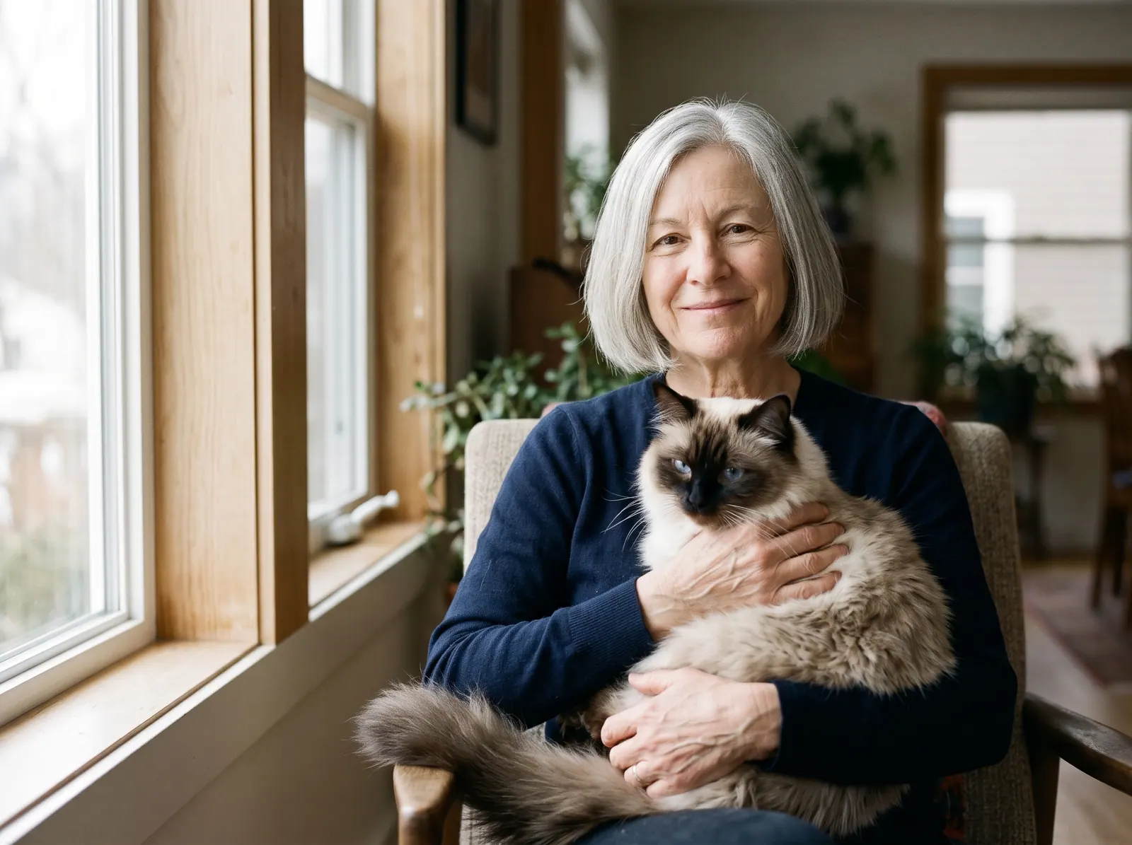 A woman with a silver bob holding her Ragdoll cat by a tall window
